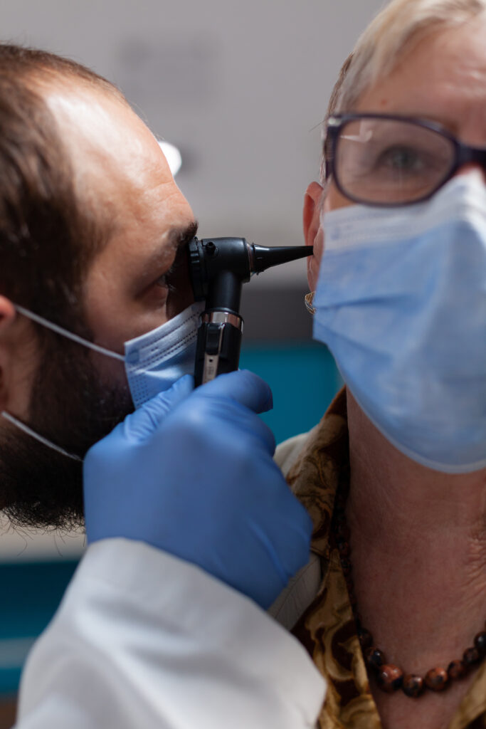 Medic holding otoscope to do ear checkup on elder woman Medic holding otoscope to do ear checkup on elder woman, wearing face mask. Physician using otolaryngology intrument at checkup consultation with patient during pandemic. Close up